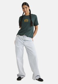 Green cotton t-shirt with yellow embroidered text, paired with light blue wide-leg jeans and black sneakers. Model poses with hands in pockets.