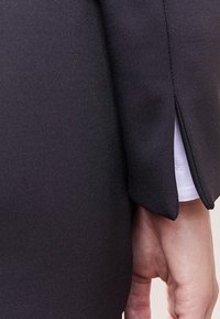 Robe noire avec un corsage ajusté et une manche légèrement évasée. Le tissu est lisse, avec une couche blanche sous-jacente au niveau du poignet de la manche.