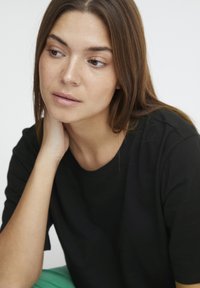 Sort bomulds T-shirt med korte ærmer. Modellen har langt, glat brunt hår og hviler sin hage på sin hånd. Glat hudstruktur.