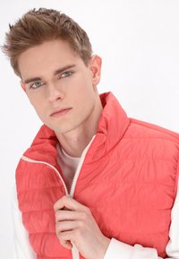 Gilet coral a trapunte con colletto alto e zip bianca, indossato sopra una camicia bianca a maniche lunghe. Texture liscia e design aderente.