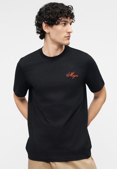 Νεαρός άνδρας με σγουρά σκούρα μαλλιά, που φοράει μαύρο μπλουζάκι MSGM και μπεζ παντελόνι, κοιτάζει προς τα δεξιά με τα χέρια πίσω από την πλάτη.