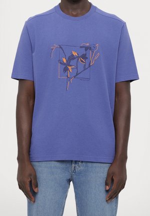 Man wearing a blue t-shirt with an abstract orange and black floral design, paired with light blue denim jeans.