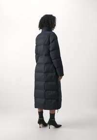 Dlouhý černý prošívaný kabát s vysokým límcem, se vzorem prošití a rovný střih. Kombinován s černými kotníkovými botami na vysokém podpatku a tmavými ponožkami.