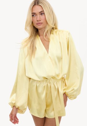 Femme blonde portant une combinaison cache-cœur à manches longues en satin jaune pâle avec une ceinture à la taille et des manches volumineuses.