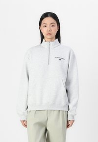 Lys grå quarter-zip hettegenser laget av hettegenserstoff med en frontlomme, og har en svart logo på brystet og elastiske mansjetter.