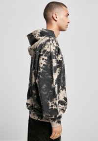Bărbat într-un hoodie negru și bej cu modele tie-dye, stând în profil, pe un fundal simplu de culoare deschisă.