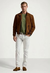 Ung man i brun mockajacka, grön skjorta, vita byxor och bruna tofflor med fransar, som står med ena handen i fickan.