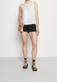 Vit ärmlös topp med spetsdetaljer och rund halsringning, kombinerad med svarta shorts och svarta sandaletter med remmar. Brun texturerad handväska.