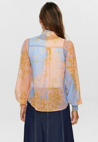 Genomskinlig blus i pastellblått och rosa med botaniska mönster. Har långa puffärmar och en volanghalsringning. Lätt och flödande tyg.