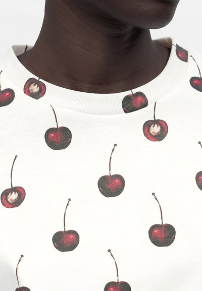 Άτομο με σκουρόχρωμο δέρμα που φοράει λευκό πουκάμισο με μοτίβο κόκκινων κερασιών με κοτσάνια, με έμφαση στον λαιμό και το πάνω μέρος του στήθους.
