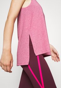 Femme portant un haut rose sans manches avec une fente latérale, associé à des leggings bordeaux présentant des rayures diagonales rose vif.