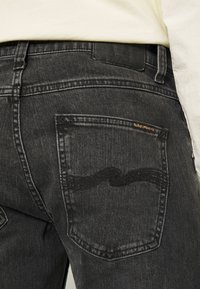 Černé džíny z denimu s zadní kapsou se vlnitým švem a malou zlatou etiketou, které předvádějí klasický střih a texturovaný materiál.