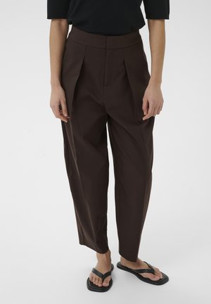 Isik, kes kannab kõrge vöökohaga tumepruune plisseeritud pükse, musta lühikeste varrukatega topi ja mustadest platvorm-sandaalidest jalanõusid, valgel taustal.