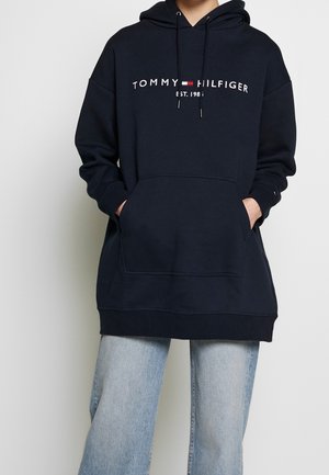 Osoba nosząca czarną bluzę z kapturem Tommy Hilfiger z przednią kieszenią i jasnoniebieskie dżinsy, ręce w kieszeniach bluzy.