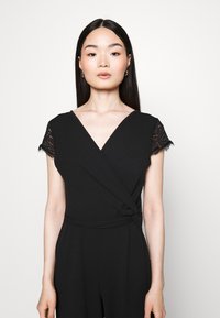 Črn kombinezon z ovitkom, ki ima kratke čipkaste rokave, V-izrez in zanko v pasu. Gladek tekstura tkanine.