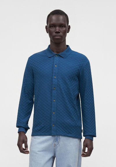 Homme portant une chemise bleue texturée à manches longues avec col et un jean bleu clair, debout devant un fond uni.