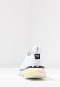 Białe buty sportowe z teksturowaną cholewką, czarnymi akcentami i kolorową gradientową podeszwą (żółta, różowa). Zawierają pętelkę z brandingiem.