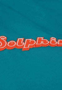 Türkiser Stoff mit besticktem "Solphin"-Logo in leuchtendem Orange. Das Logo hat eine markante, verspielte Schriftart mit einer leichten 3D-Textur.