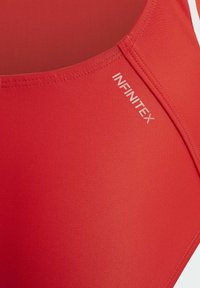 Bañador rojo hecho de un material suave y elástico con detalles en blanco. Presenta el logotipo "INFINITEX" y un diseño aerodinámico y atlético.