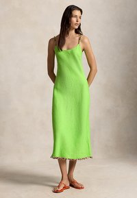 Nő áll, élénk zöld, ujjatlan midiruha van rajta, színes gyöngyös pántokkal és szegéllyel, narancssárga szandállal párosítva.