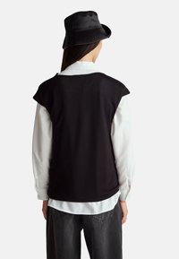 Černý bezramenný top s hladkou texturou, vrstvený přes bílou košili s dlouhým rukávem. Model má na sobě černý klobouk typu bucket a tmavé džíny.