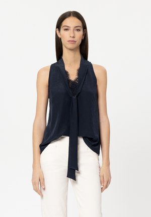 Marineblaue ärmellose Bluse mit Vorderbindeband und Spitzenbesatz am Ausschnitt, aus einem glatten Stoff gefertigt. Kombiniert mit hellen Hosen.