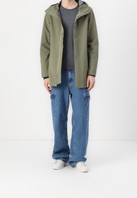 Vojensky zelená nepromokavá bunda s kapucí, zipovým zapínáním a barevným akcentem. Kombinována s volnými modrými denimovými cargo kalhotami a bílými teniskami.