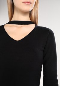 Svart ribbad stickad tröja med V-ringning och ett horisontellt utskärningsdetalj vid den  övre halsringningen. Långa ärmar och figurnära design.