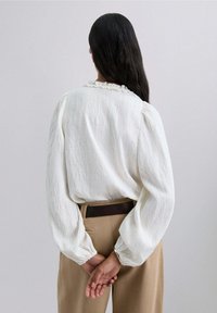 Blusa blanca de textura con mangas abullonadas, que presenta un cuello con volante, combinada con pantalones beiges de cintura alta y un cinturón marrón.