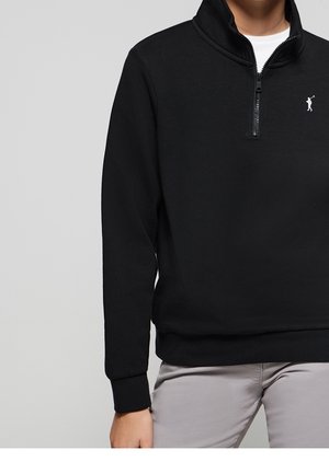 Person iført en sort quarter-zip pullover med et lille hvidt golfer-logo på brystet og lysegrå bukser mod en ensfarvet baggrund.