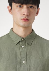 Camicia collare verde oliva realizzata in tessuto strutturato, con chiusura a bottoni frontale e logo "Lee" ricamato sul petto.