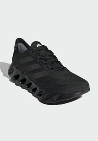 Black athletic shoe with a mesh upper, three black stripes, and a textured black sole featuring elliptical cutouts for flexibility.