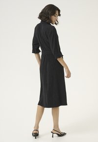 Robe noire en tissu texturé avec une taille ceinturée et des manches ¾. Le design présente des détails plissés et un col classique. Ourlet évasé.