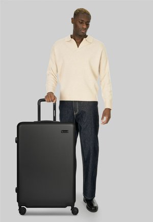 Mann in cremefarbenem Pullover und dunkler Jeans stehend, hält schwarzen Rollkoffer mit ausgezogenem Griff vor grauem Hintergrund.