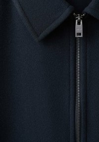 Granatowa kurtka z mieszanki wełny z kołnierzem w szpic i srebrnym metalowym zamkiem błyskawicznym. Charakteryzuje się gładką fakturą i dopasowanym krojem.