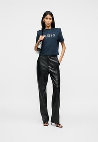 Γυναίκα με μπλε μπλούζα Guess, μαύρο δερμάτινο παντελόνι ψηλής μέσης, μαύρα σανδάλια με τακούνι και μαύρη τσάντα ώμου.