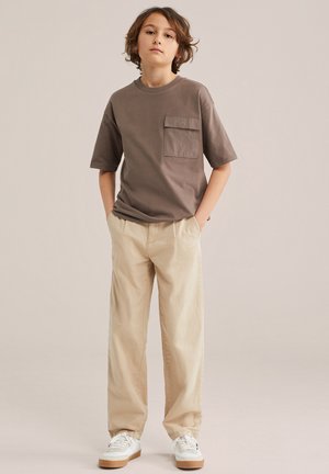 Keskmise pikkusega pruunide juustega poiss, kes kannab pruuni taskutega T-särki, beeže pükse ja valgeid tossusid, seistes käed taskutes.