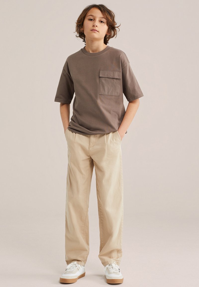 Garçon aux cheveux bruns mi-longs portant un t-shirt marron avec poches, un pantalon beige et des baskets blanches, debout les mains dans les poches.