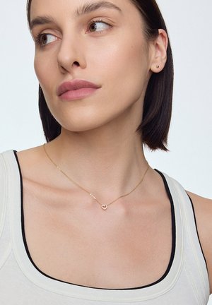 Złoty naszyjnik z zawieszką w kształcie serca, z małymi koralikami na delikatnym łańcuszku, noszony przez osobę o krótkich ciemnych włosach, na jasnym tle.