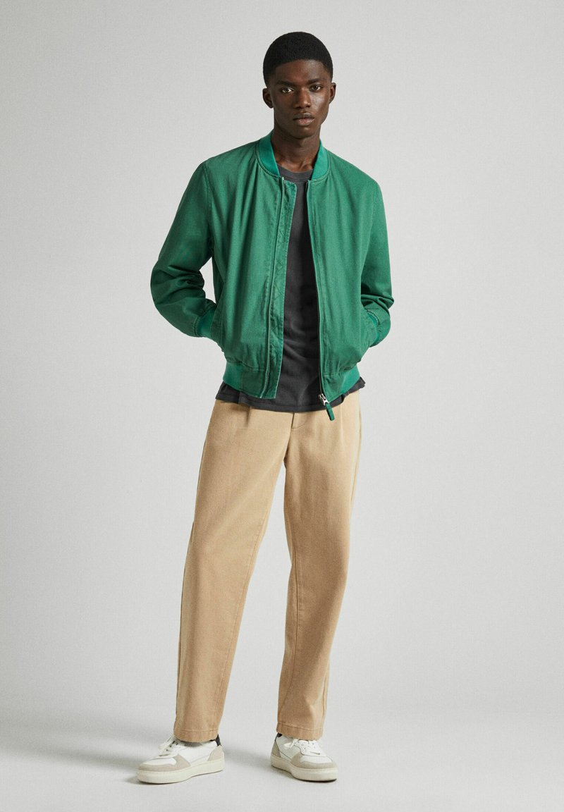 Chaqueta bomber verde con cremallera, combinada con pantalones beige y zapatillas blancas. La chaqueta presenta un cuello acanalado y un ajuste ligeramente holgado.