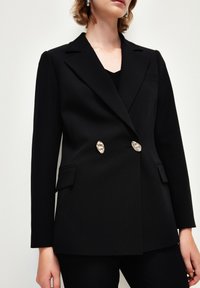 Černý přizpůsobený blazer vyrobený z hladké tkaniny. Disponuje dvouřadým designem, stříbrnými knoflíky a strukturovanými rameny s kapsami.