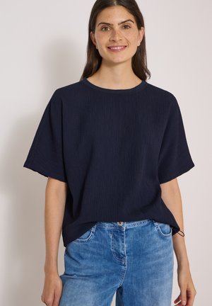 Frau mit langen dunklen Haaren und Sommersprossen, die ein locker sitzendes, dunkel marineblaues, strukturiertes Kurzarmshirt und hellblaue Jeans trägt, lächelt in die Kamera.