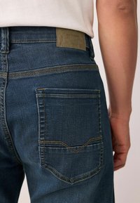 Sötétkék farmernadrág, enyhén kopott textúrával, látható varrással és jellegzetes hátsó zsebtervezéssel, logó címkével.