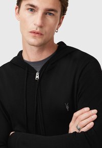 Sudadera negra con cremallera y cordón, que presenta un logo gris bordado en el pecho. Textura suave y diseño ajustado.