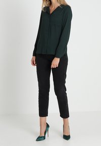 Blusa verde scuro con colletto, abbottonatura frontale e taschino sul petto, abbinata a pantaloni neri a sigaretta e scarpe con tacco teal.