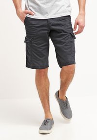 Hombre que lleva pantalones cortos de carga gris oscuro, camiseta gris claro y zapatillas de lona grises, de pie con una mano en el bolsillo.