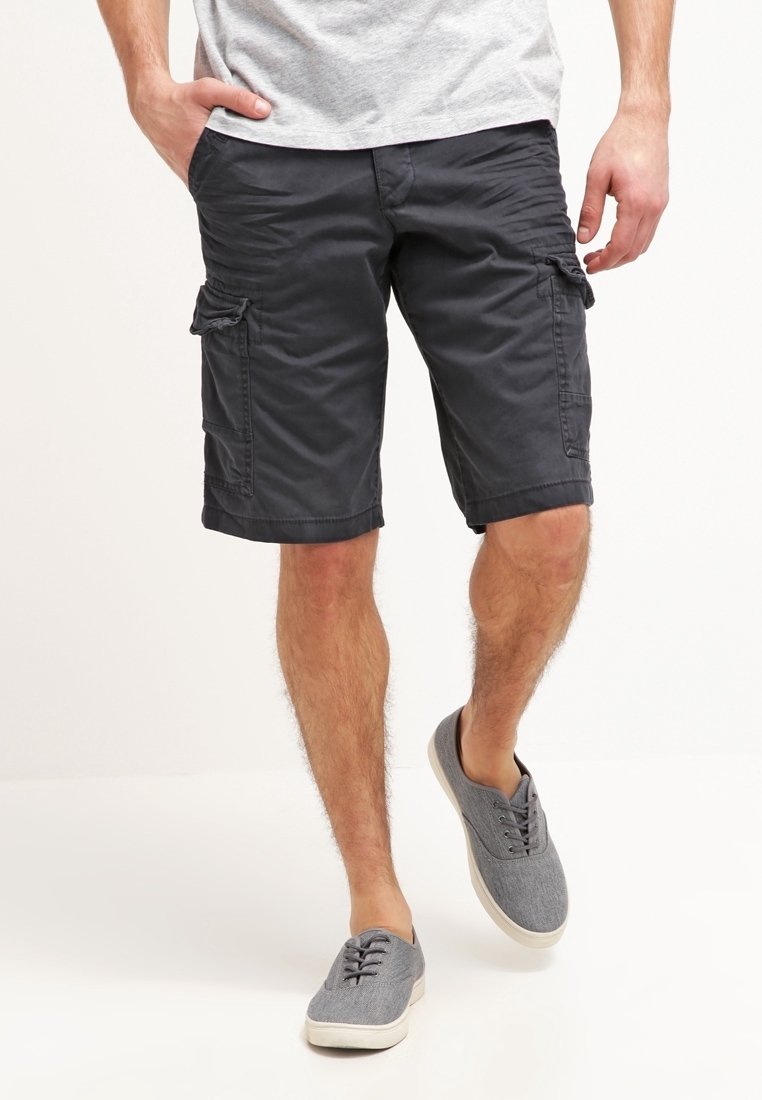 Hombre que lleva pantalones cortos de carga gris oscuro, camiseta gris claro y zapatillas de lona grises, de pie con una mano en el bolsillo.