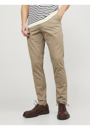 Man in beige chino's, bruine leren veterschoenen en een wit met marineblauwe gestreepte trui, staande met handen in de zakken.