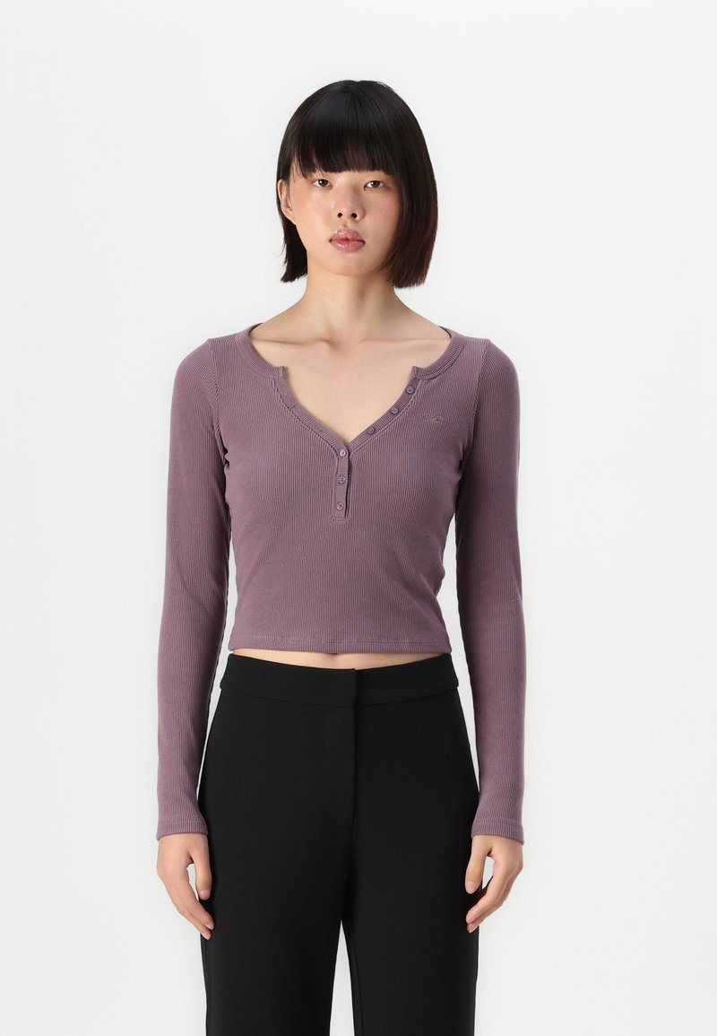 Hosszú ujjú, roppantott anyagból készült crop top, mély V-kivágással és elől gombos záródással, tompa lila színben. Fekete nadrággal párosítva.