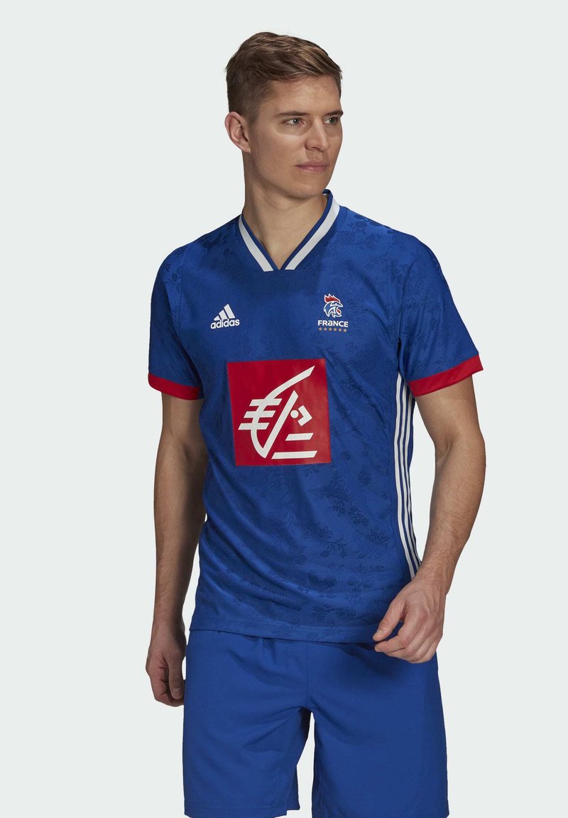 Jonge man in een blauwe Frankrijk voetbalshirt met rode en witte accenten, Adidas-logo en sponsor-embleem, die opzij kijkt.